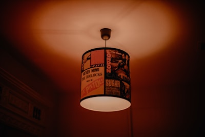A ceiling light with a lampshade decorated with vintage rock band posters and album covers. The ambient light creates a warm glow, casting soft shadows on the ceiling.