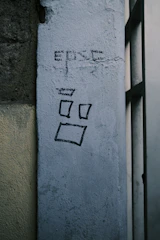 A moody, textured urban wall with faded graffiti in washed black and sand tones.