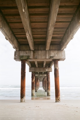 A perspective view under a pier with wooden beams and concrete pillars stretching into the distance. The metal pillars show signs of rust and are partially submerged in water. The ocean waves are visible to the sides, and the sandy beach lies in the foreground.