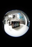 A fisheye lens view showcasing a set of Testuggine brand vape devices in various colors, displayed on a white table. The background features modern pendant lights and horizontal blinds with reflections, adding a futuristic and sleek ambiance.