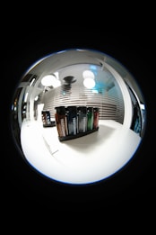 A fisheye lens view showcasing a set of Testuggine brand vape devices in various colors, displayed on a white table. The background features modern pendant lights and horizontal blinds with reflections, adding a futuristic and sleek ambiance.