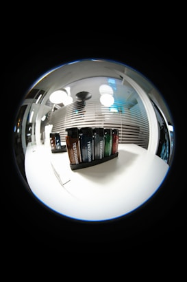 A fisheye lens view showcasing a set of Testuggine brand vape devices in various colors, displayed on a white table. The background features modern pendant lights and horizontal blinds with reflections, adding a futuristic and sleek ambiance.