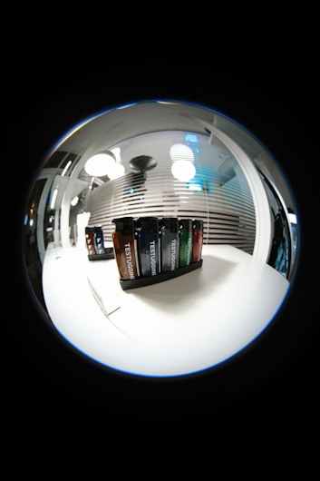A fisheye lens view showcasing a set of Testuggine brand vape devices in various colors, displayed on a white table. The background features modern pendant lights and horizontal blinds with reflections, adding a futuristic and sleek ambiance.
