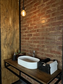 A modern bathroom vanity setup featuring a rectangular white sink on a wooden countertop. Above the sink, an exposed bulb hanging light fixture adds a rustic touch. The surrounding brick wall enhances the cozy, industrial feel. On the counter, there's a small plant, a soap dispenser, and a black wicker basket holding paper towels.