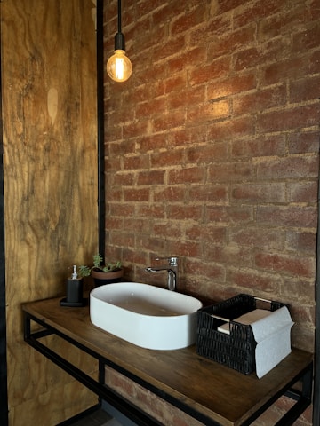 A modern bathroom vanity setup featuring a rectangular white sink on a wooden countertop. Above the sink, an exposed bulb hanging light fixture adds a rustic touch. The surrounding brick wall enhances the cozy, industrial feel. On the counter, there's a small plant, a soap dispenser, and a black wicker basket holding paper towels.