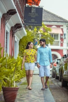 A couple walks hand in hand on a sidewalk lined with potted plants. They are smiling at each other, dressed in casual, colorful outfits. The building beside them is painted red with white trim, featuring wrought iron details and a sign for Panjim Inn. Cars are parked along the street, and the setting conveys a relaxed, sunny day.