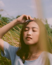 A person with long hair stands outdoors with their eyes closed, lightly touching their forehead. Surrounding them are tall grasses, and their expression appears peaceful. A soft, natural light creates a calming atmosphere.