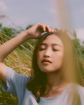 A person with long hair stands outdoors with their eyes closed, lightly touching their forehead. Surrounding them are tall grasses, and their expression appears peaceful. A soft, natural light creates a calming atmosphere.