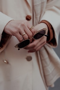 A close-up of hands holding a pair of sunglasses. One hand is adorned with gold rings, adding a touch of elegance to the scene. The coat buttoned in the background suggests a sophisticated fashion style.