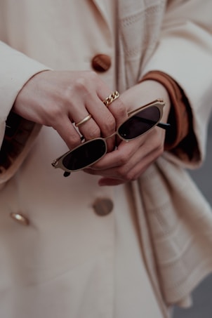 A close-up of hands holding a pair of sunglasses. One hand is adorned with gold rings, adding a touch of elegance to the scene. The coat buttoned in the background suggests a sophisticated fashion style.