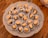 A decorative plate holds several small confections, each placed in a white paper cup. These treats consist of a light caramel-colored filling, likely a nut, set atop a dried fruit, possibly a prune or date. The plate rests on a wooden surface, adding a rustic touch.