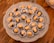 A decorative plate holds several small confections, each placed in a white paper cup. These treats consist of a light caramel-colored filling, likely a nut, set atop a dried fruit, possibly a prune or date. The plate rests on a wooden surface, adding a rustic touch.