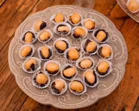 A decorative plate holds several small confections, each placed in a white paper cup. These treats consist of a light caramel-colored filling, likely a nut, set atop a dried fruit, possibly a prune or date. The plate rests on a wooden surface, adding a rustic touch.
