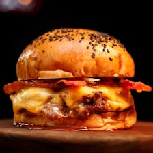 A juicy hamburger with a toasted bun topped with sesame seeds. Melting cheese oozes over a beef patty, complemented by crispy slices of bacon. The burger sits on a wooden surface with a dark, blurred background.