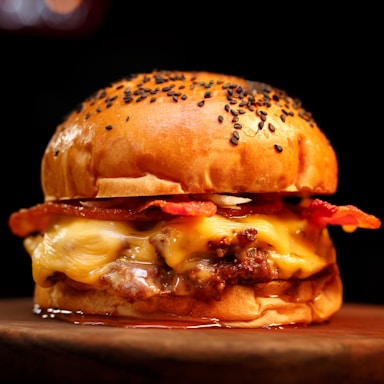 A juicy hamburger with a toasted bun topped with sesame seeds. Melting cheese oozes over a beef patty, complemented by crispy slices of bacon. The burger sits on a wooden surface with a dark, blurred background.