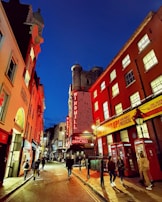 A narrow street at night flanked by buildings with vibrant neon lights, including a prominent sign for a venue called Windmill. The street features classic British red telephone booths and several people walking along the sidewalk. The sky is a deep blue, and the illumination from the signs creates a warm glow on the surrounding structures.