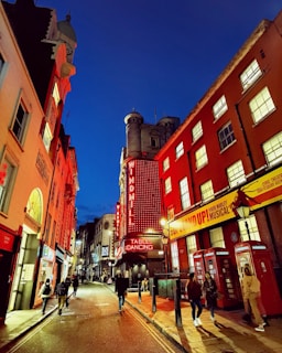 A narrow street at night flanked by buildings with vibrant neon lights, including a prominent sign for a venue called Windmill. The street features classic British red telephone booths and several people walking along the sidewalk. The sky is a deep blue, and the illumination from the signs creates a warm glow on the surrounding structures.