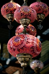 Ornate hanging lamps with intricate mosaic patterns in shades of red, pink, and gold. The lamps have a vintage look with bronze metalwork and colorful glass pieces creating geometric designs. They are suspended in an indoor setting, providing an elegant and warm ambiance.