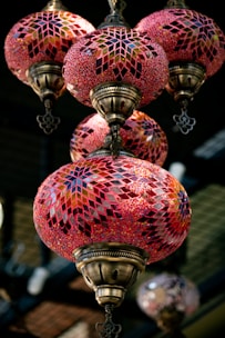 Ornate hanging lamps with intricate mosaic patterns in shades of red, pink, and gold. The lamps have a vintage look with bronze metalwork and colorful glass pieces creating geometric designs. They are suspended in an indoor setting, providing an elegant and warm ambiance.
