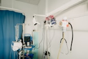 A hospital room with medical equipment, including an IV stand, a blood pressure monitor, a suction canister, and various medical instruments attached to a wall. The room features a blue privacy curtain and a shelf with medical supplies.