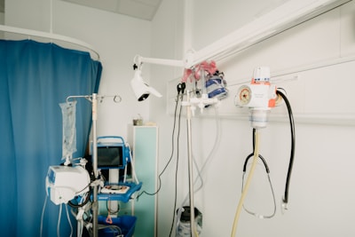 A hospital room with medical equipment, including an IV stand, a blood pressure monitor, a suction canister, and various medical instruments attached to a wall. The room features a blue privacy curtain and a shelf with medical supplies.