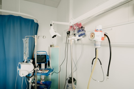 A hospital room with medical equipment, including an IV stand, a blood pressure monitor, a suction canister, and various medical instruments attached to a wall. The room features a blue privacy curtain and a shelf with medical supplies.