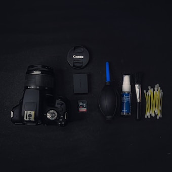 A black Canon camera with a zoom lens is placed on a dark surface alongside various camera accessories. These accessories include a lens cap, a camera battery, a SanDisk memory card, a rubber air blower, a bottle of cleaning fluid, a cleaning brush, and several cotton swabs arranged neatly.