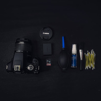 A black Canon camera with a zoom lens is placed on a dark surface alongside various camera accessories. These accessories include a lens cap, a camera battery, a SanDisk memory card, a rubber air blower, a bottle of cleaning fluid, a cleaning brush, and several cotton swabs arranged neatly.