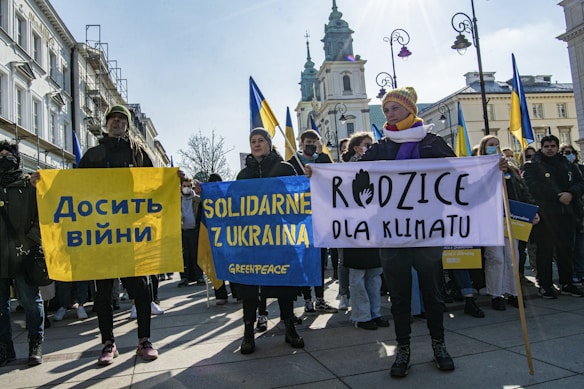A group of people is gathered on a city street holding banners with messages of support for Ukraine and climate action. The banners display texts in different languages, including Ukrainian and Polish. The scene is set in front of historical buildings with clear skies in the background.