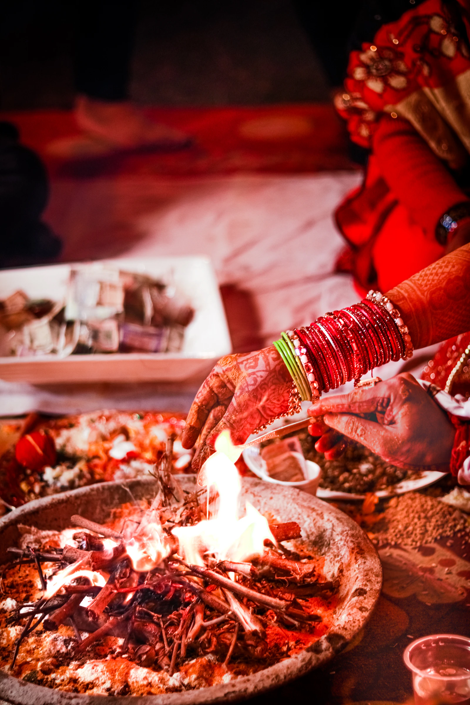 चूड़ियों और मेहंदी वाला हाथ हवन कुंड में आहुति डालते हुए, विवाह की अग्नि के सामने।