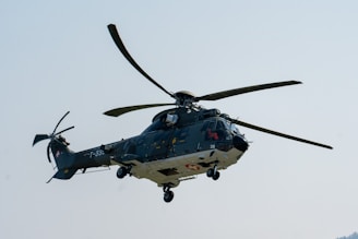 A military helicopter is captured mid-flight against a clear blue sky. The helicopter features a green and grey color scheme, with the tail rotor visible and landing gear extended. Distinctive markings, including a red cross on a white background and alphanumeric codes, are present on the body of the aircraft.