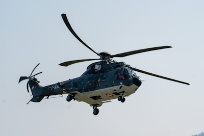 A military helicopter is captured mid-flight against a clear blue sky. The helicopter features a green and grey color scheme, with the tail rotor visible and landing gear extended. Distinctive markings, including a red cross on a white background and alphanumeric codes, are present on the body of the aircraft.