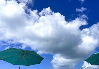 Two teal umbrellas are positioned at the bottom corners against a backdrop of vibrant blue sky and large, fluffy white clouds.