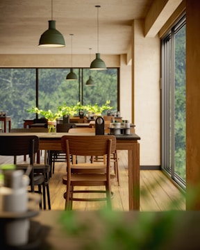 A bright and airy dining area features rows of wooden tables and chairs. Large windows allow natural light to flood the space, offering a view of lush greenery outside. Green pendant lights hang from the ceiling, and small vases with green plants adorn the tables.