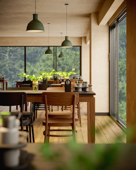 A bright and airy dining area features rows of wooden tables and chairs. Large windows allow natural light to flood the space, offering a view of lush greenery outside. Green pendant lights hang from the ceiling, and small vases with green plants adorn the tables.