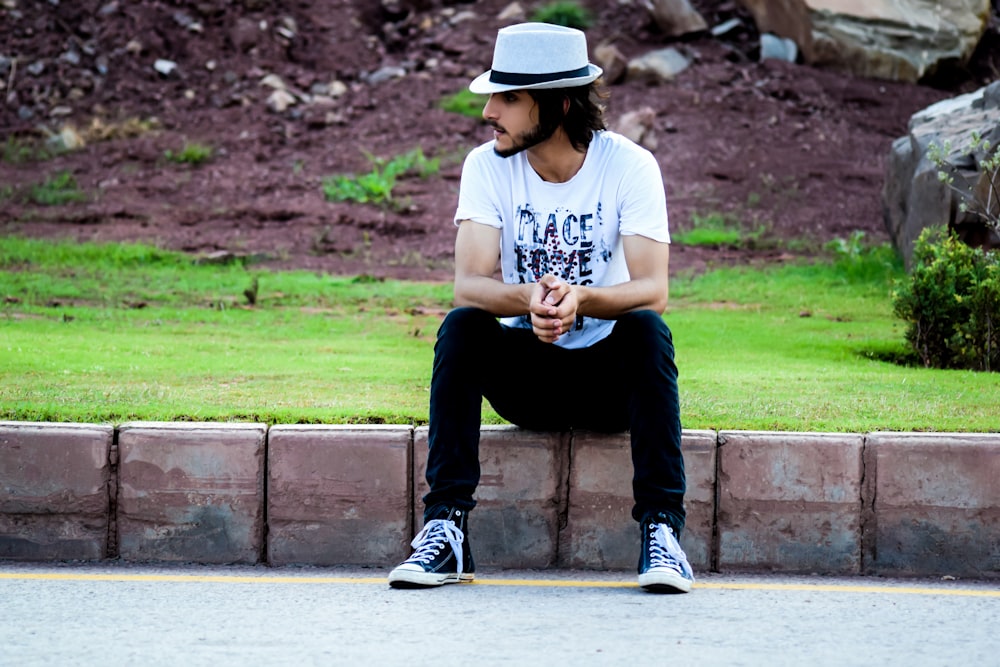 a man sitting on the side of a road wearing a hat