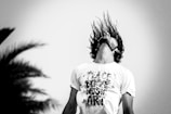 A black-and-white image featuring a person with long hair flipping their head back, causing their hair to fly upward. They are wearing a white T-shirt with text that reads 'PEACE LOVE MUSIC ART.' The background includes an out-of-focus silhouette of a tree or plant.