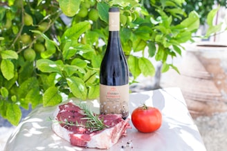 A bottle of red wine is placed on a white surface alongside a raw beef steak garnished with rosemary and a ripe tomato. In the background, there are lush green leaves, likely from a lemon or lime tree.