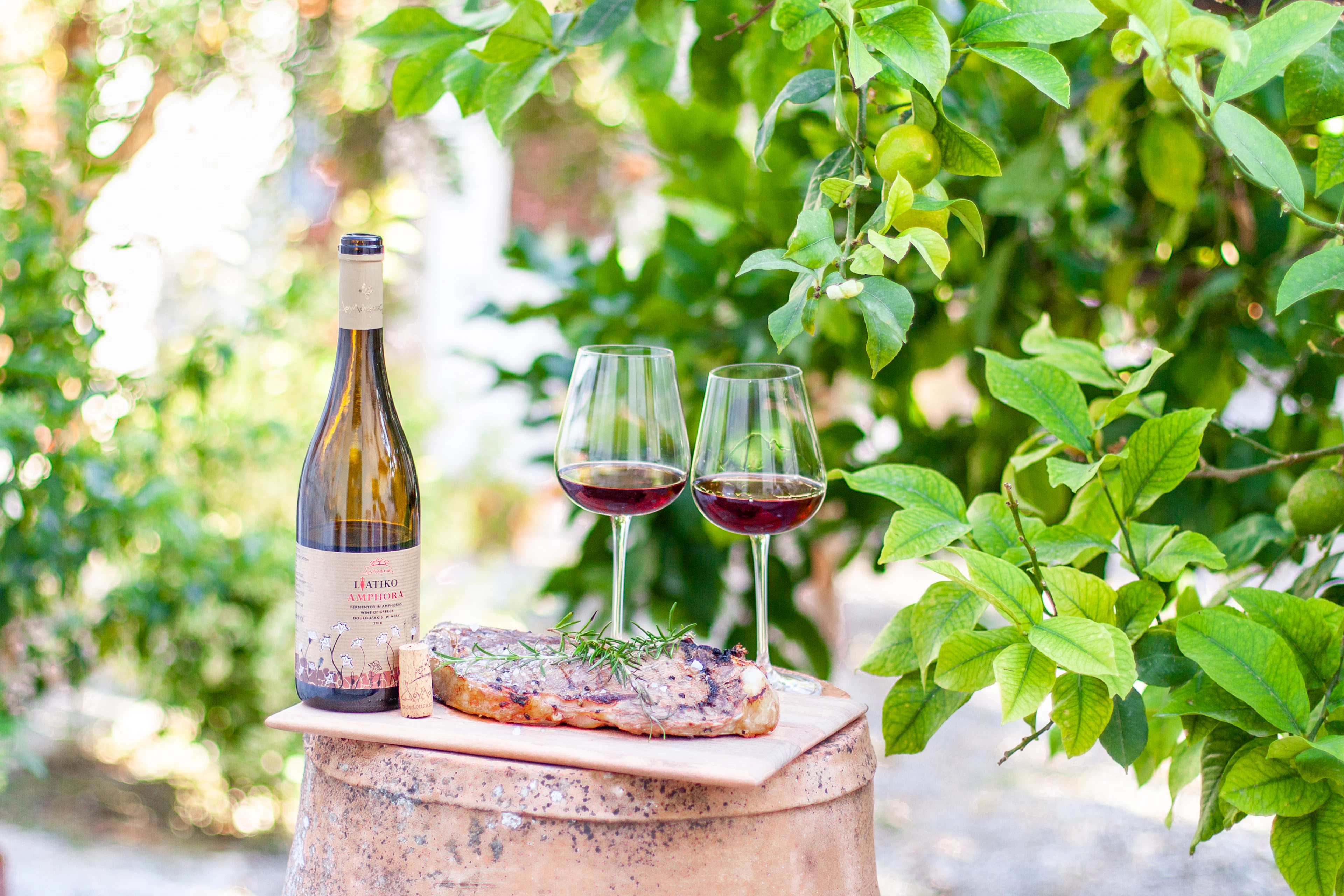 A bottle of wine, two filled glasses and grilled meat are arranged on a barrel in a leafy garden.