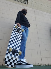 A person wearing a black face mask, dark hoodie, and blue jeans holds a black-and-white checkered skateboard. The photo is taken from a low angle, highlighting the skateboard and the person's casual shoes. The background shows a textured brick wall.