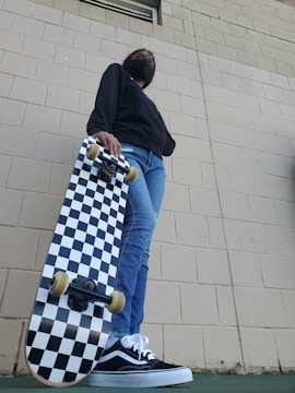 A person wearing a black face mask, dark hoodie, and blue jeans holds a black-and-white checkered skateboard. The photo is taken from a low angle, highlighting the skateboard and the person's casual shoes. The background shows a textured brick wall.