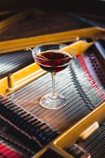 A cocktail glass filled with a dark red beverage is placed on the strings of a piano. The piano's interior is visible with its metallic strings and wooden frame, showcasing the intricate details of its construction. The light reflects on the glass and liquid, creating a bright contrast against the muted backdrop of the piano.