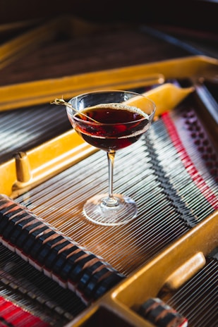 A cocktail glass filled with a dark red beverage is placed on the strings of a piano. The piano's interior is visible with its metallic strings and wooden frame, showcasing the intricate details of its construction. The light reflects on the glass and liquid, creating a bright contrast against the muted backdrop of the piano.