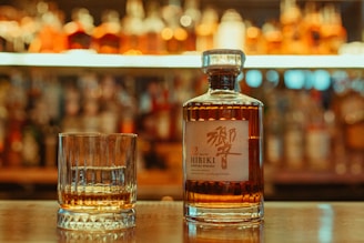 A bottle of Hibiki Japanese whisky and a glass with a small amount of whisky are placed on a bar counter. The background features an array of blurred liquor bottles, suggesting a warm, ambient bar setting.