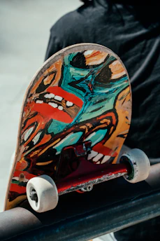 A close-up view of the underside of a skateboard, featuring vibrant and abstract artwork with bold colors such as turquoise, red, and yellow. The skateboard's wheels and trucks are visible, resting on a smooth metal surface. A person, partially visible, wearing a dark jacket is in the background.