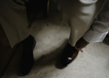 A person in formal attire is adjusting or wearing a brown leather shoe. The setting appears to be indoors, with a focus on well-tailored trousers and a white long-sleeved shirt.