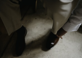 A person in formal attire is adjusting or wearing a brown leather shoe. The setting appears to be indoors, with a focus on well-tailored trousers and a white long-sleeved shirt.