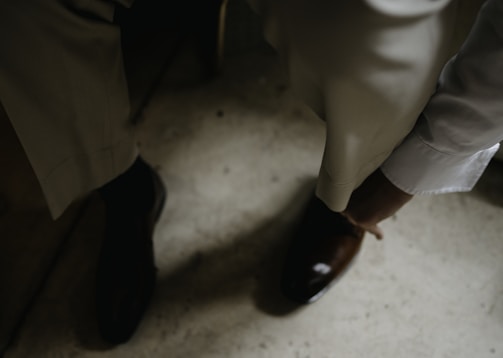 A person in formal attire is adjusting or wearing a brown leather shoe. The setting appears to be indoors, with a focus on well-tailored trousers and a white long-sleeved shirt.