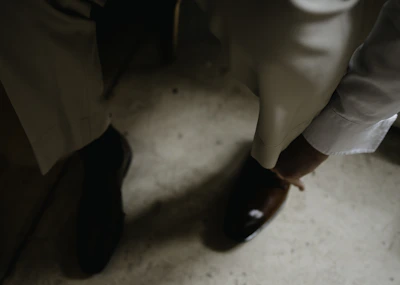 A person in formal attire is adjusting or wearing a brown leather shoe. The setting appears to be indoors, with a focus on well-tailored trousers and a white long-sleeved shirt.
