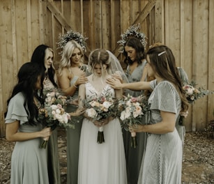 A group of women in elegant dresses surround a bride, gently placing their hands on her shoulders and back. Each woman holds a bouquet of flowers with pastel shades of pink, peach, and white. The setting appears rustic with a wooden backdrop, and two circular floral arrangements hang on the wall behind them.